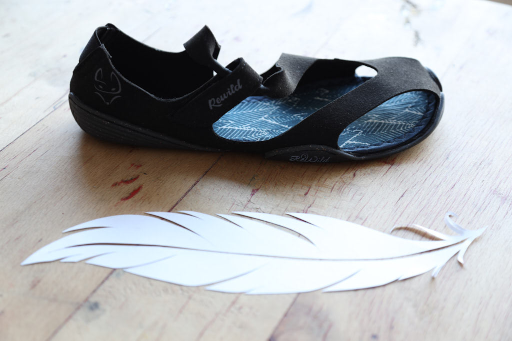 Eine schwarze Wildling Shoes Sandale auf Holszdielen, daneben eine große weiße Feder.