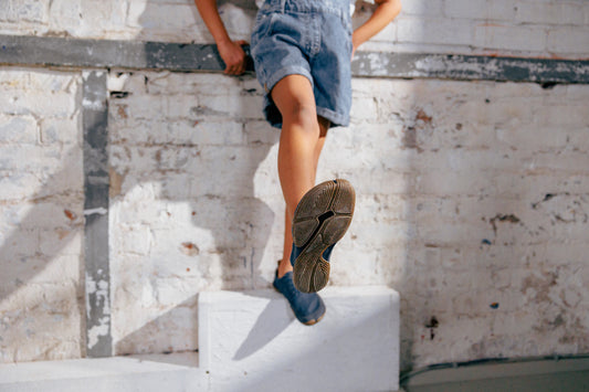 Vor einem hellen Mauerwerk steht auf einem niedrigen, hellen Quader eine Person in kurzen Jeans und Wildling Minimalschuhen. Die Person ist bis zur Hüfte im Bild und streckt ein Bein nach vorne, sodass die Sohle eines Schuhs Richtung Kamera gedreht ist.