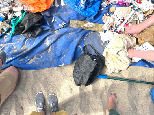 Ein Strand, darauf ist eine blaue Decke ausgebreitet, auf der gesammelter Müll abgelegt wurde. Am unteren Bildrand sind die Füße von drei Personen zu sehen, am rechten Bildrand sieht man zwei Hände.