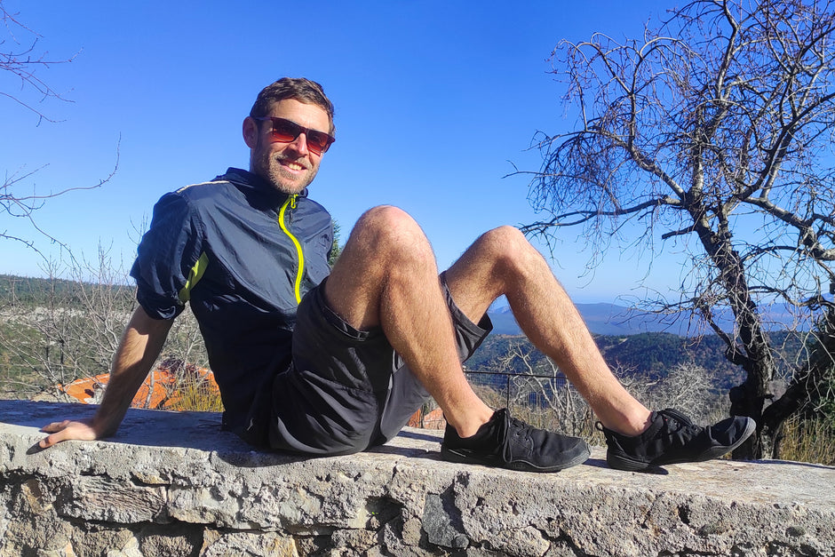 Marco von Reisefroh sitzt auf einer Mauer und lächelt Richtung Kamera. Er trägt sommerliche Outdoor-Kleidung, eine Sonnenbrille und schwarze Wildlinge. Im Hintergrund blauer Himmel und Landschaft.