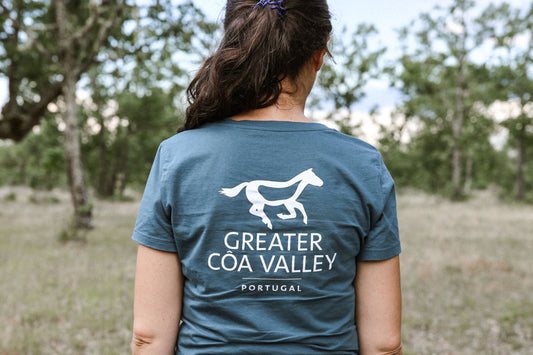 Eine Person in einer grünen Landschaft von hinten in einem petrolfarbenen T-Shirt mit dem Aufdruck "Greater Côa Valley Portugal".
