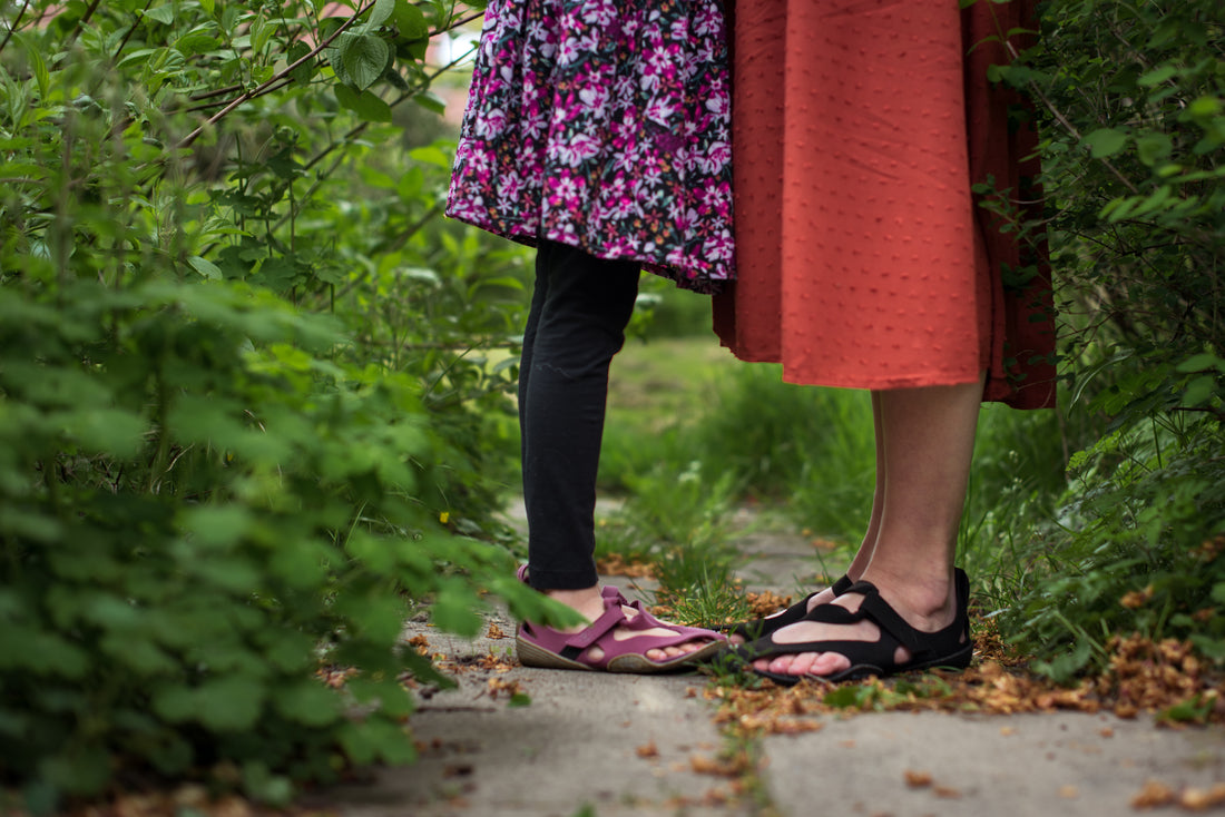 Zwei Personen bis etwa zur Hüfte. Sie stehen auf einem schmalen Weg zwischen Büsche, tragen farbige Röcke und Wildling Barfußschuhe.