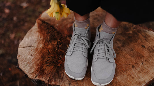 Nahaufnahme: Zwei Füße in grauen Wildling Schuhen stehen auf einem Baumstumpf. Am Bildrand gelbe Blumen.