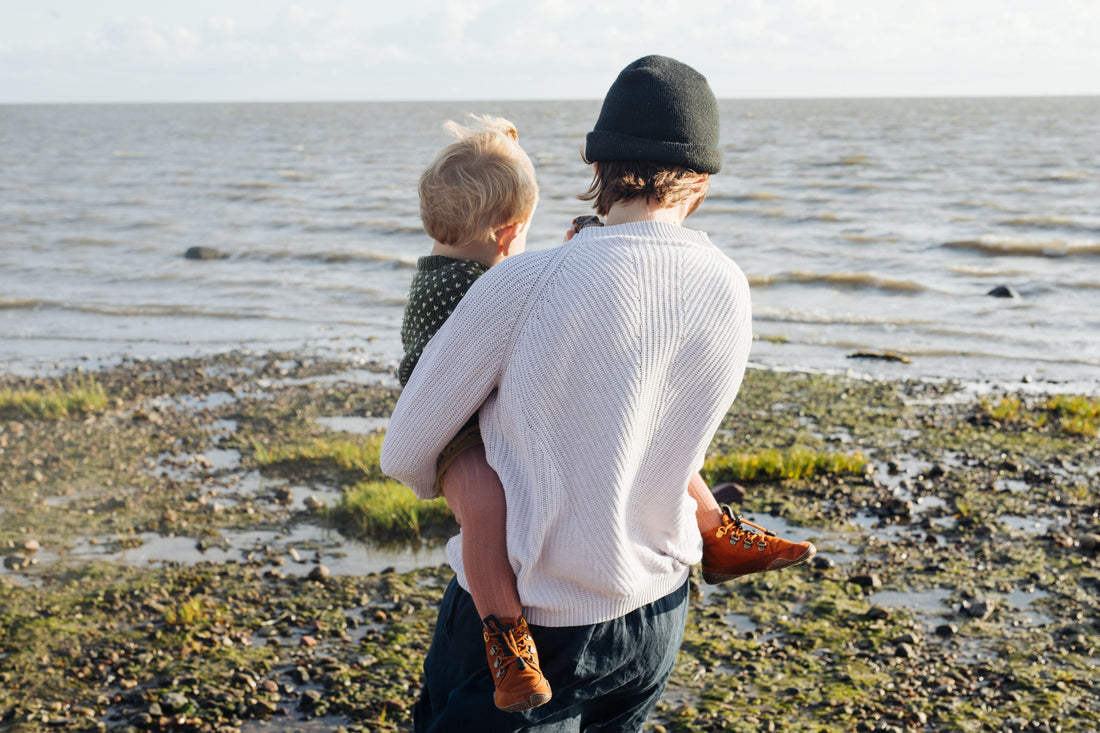 Eine erwachsene Personen, ein Kind auf dem Arm haltend, von hinten aufgenommen. Die Person steht auf einem steinigen Strand und hat den Blick aufs Meer gerichtet.