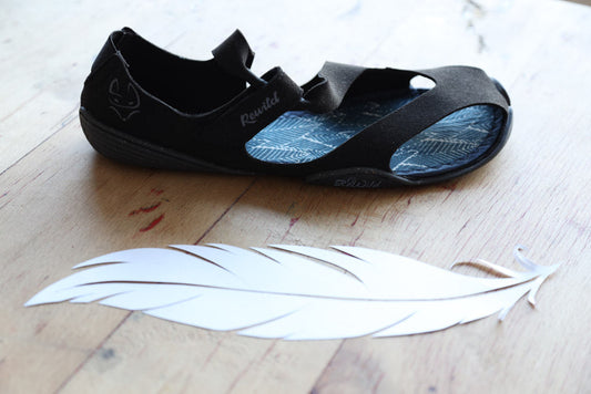Eine schwarze Wildling Shoes Sandale auf Holszdielen, daneben eine große weiße Feder.