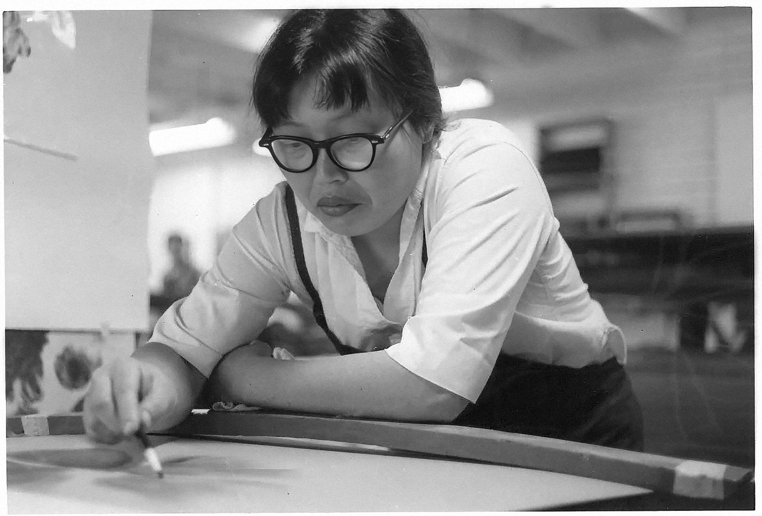 Die Künstlerin Ruth Asawa konzentriert bei der Arbeit: Sie ist über einen Tisch gebeugt und zeichnet ein neues Kunstwerk in ihrem Studio.