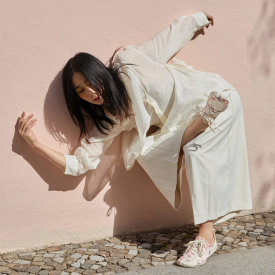 Dynamische Tanz-Pose an einer rosa Wand: Eine Frau im weißen Outfit präsentiert die gemusterten Barfußschuhe Wildling Tanuki Asawa bei einer Bewegung im Freien.